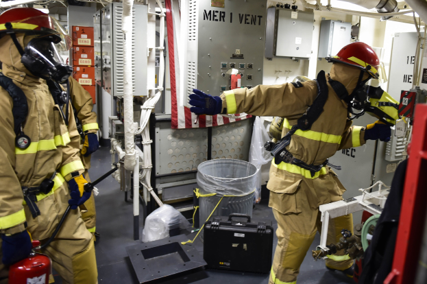 Drei Feuerwehrleute in Schutzausrüstung stehen zusammen in einem Raum mit einem Feuerlöscher links, einer Maschine rechts und verschiedenen Gegenständen und Rohren im Hintergrund.