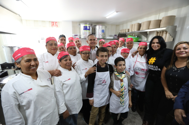 Eine Gruppe von Menschen in einer Küche, die fr├Âhlich in die Kamera l├Ąchelt und posiert, einige tragen Mützen, mit Utensilien auf der linken Seite, Regalen mit Geschirr und Deckenleuchten im Hintergrund, was auf eine Feier in einer Kochschule in Mexiko hindeutet.