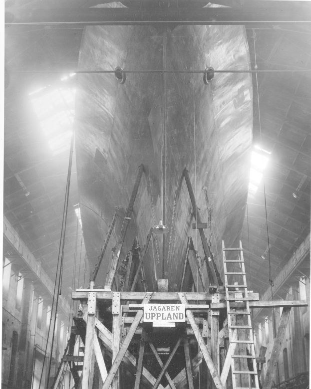 Schwarzes und weißes Foto eines großen Bootes im Bau in einer Werft, das eine Leiter, Holzpfosten, ein Schild, Metallrahmen, Deckenleuchten und Fenster zeigt.