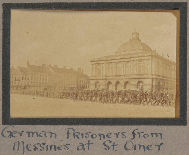 Eine Gruppe deutscher Gefangener aus Messines vor einem Gebäude unter einem klaren blauen Himmel, mit Text am unteren Bildrand.