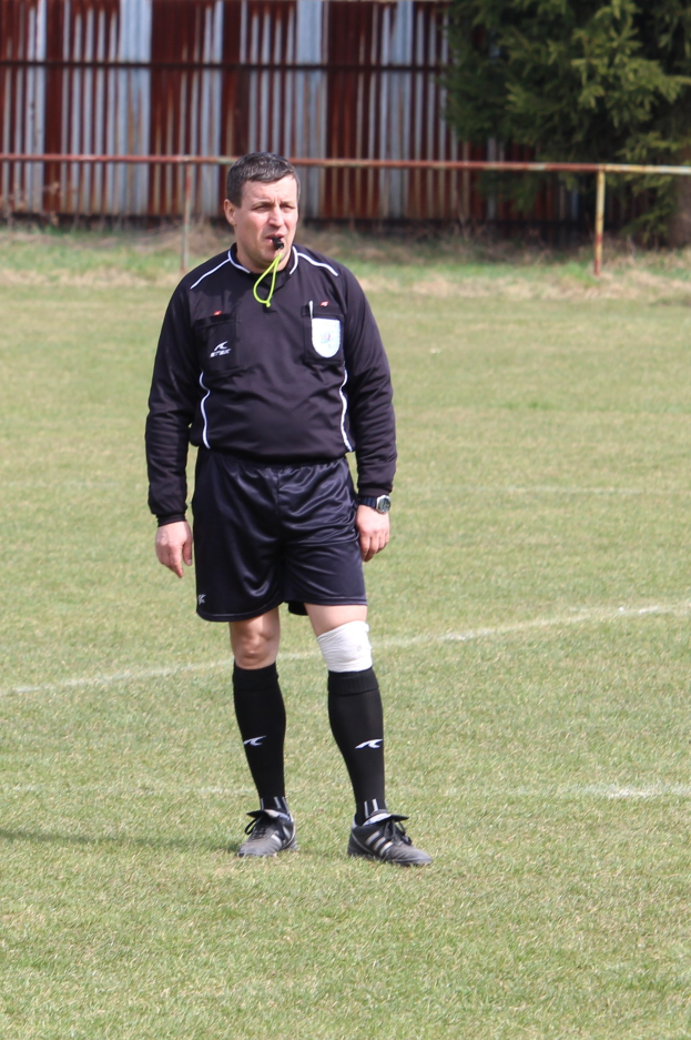 A soccer referee in uniform stands on a field surrounded by a fence and trees.