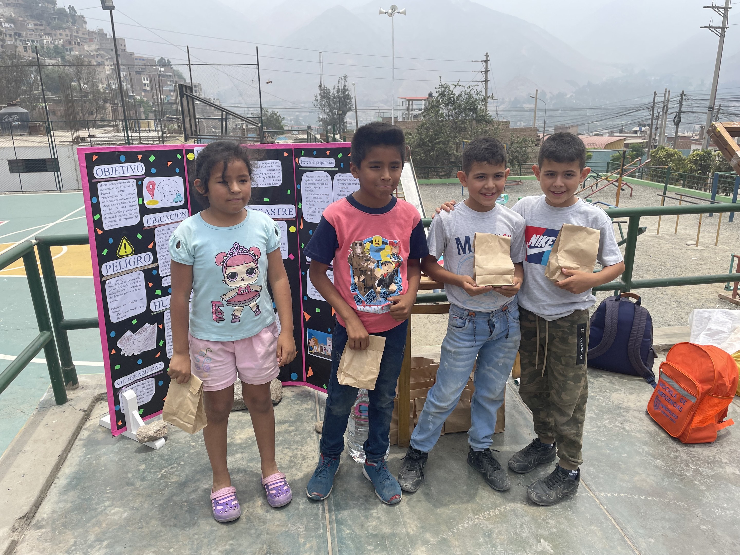 Eine Gruppe von Kindern hält Papiertüten in den Händen und steht auf einem Basketballplatz, rechts von ihnen liegen Tüten und Gegenstände auf dem Boden und links eine Tafel mit Text, im Hintergrund Zäune, Pfosten, Laternen, Strommasten, Bäume, Gebäude, Hügel und Himmel.