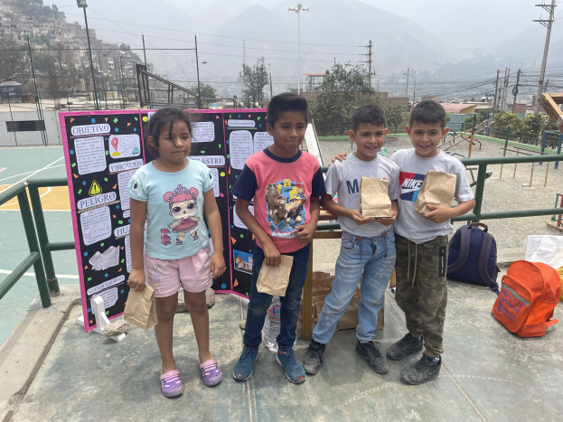 Eine Gruppe von Kindern hält Papiertüten in den Händen und steht auf einem Basketballplatz, rechts von ihnen liegen Tüten und Gegenstände auf dem Boden und links eine Tafel mit Text, im Hintergrund Zäune, Pfosten, Laternen, Strommasten, Bäume, Gebäude, Hügel und Himmel.
