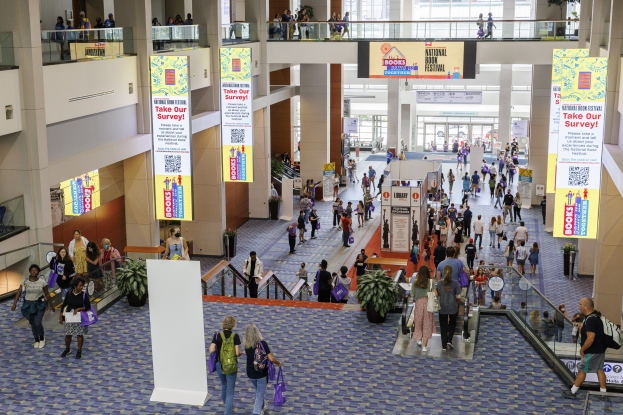 Eine große Menschenmenge, die durch ein großes Gebäude mit Bannern, Topfpflanzen, Rolltreppen, Geländern, Säulen, Texttafeln, Glastüren und anderen Gegenständen geht, wobei einige Taschen tragen, auf dem Nationalen Buchfest.