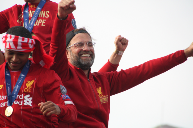 Liverpool-Trainer Jürgen Klopp feiert mit seinen Spielern den Sieg der UEFA Champions League, während zwei medaillentragende Personen in der Nähe sind und der Himmel im Hintergrund zu sehen ist.