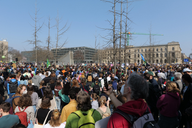 Eine große Menschenmenge nimmt an einer Klimawandel-Demonstration teil, hält Schilder und trägt Taschen, steht vor einem Gebäude mit Fenstern und Bäumen unter einem klaren Himmel.