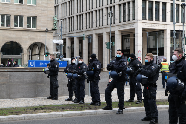 Polizeibeamte in schwarzen Uniformen und Masken stehen vor einem Berliner Gebäude mit Glasfenstern und Säulen, einige halten Helme, mit Laternenmasten, Verkehrszeichen, Schildern mit Text, einer Statue und anderen Menschen im Hintergrund.