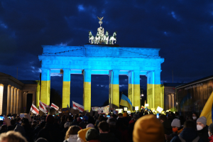 Menschenmenge mit Fahnen und Schildern vor dem Brandenburger Tor in Berlin, mit einer Banner auf der rechten Seite des Bildes.