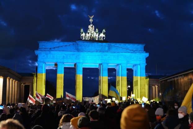 Menschenmenge mit Fahnen und Schildern vor dem Brandenburger Tor in Berlin, mit einer Banner auf der rechten Seite des Bildes.