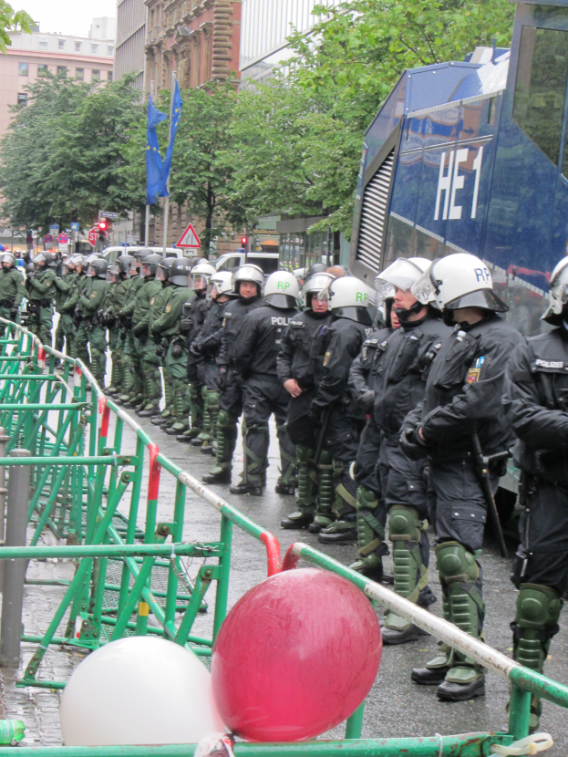 Eine Gruppe von Polizisten in Helmen und mit Waffen vor einer Absperrung auf einer Straße mit Geländern und Luftballons, mit Bäumen, Gebäuden, Flaggen und einem klaren blauen Himmel im Hintergrund.