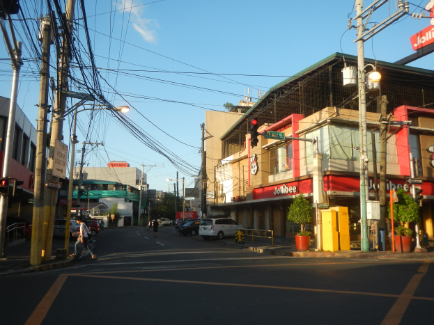 Eine belebte Stadtstraße mit Autos, Fußgängern, Strommasten, Laternen, Ampeln, Gebäuden mit Schildern, Topfpflanzen und einer bewölkten Himmel.