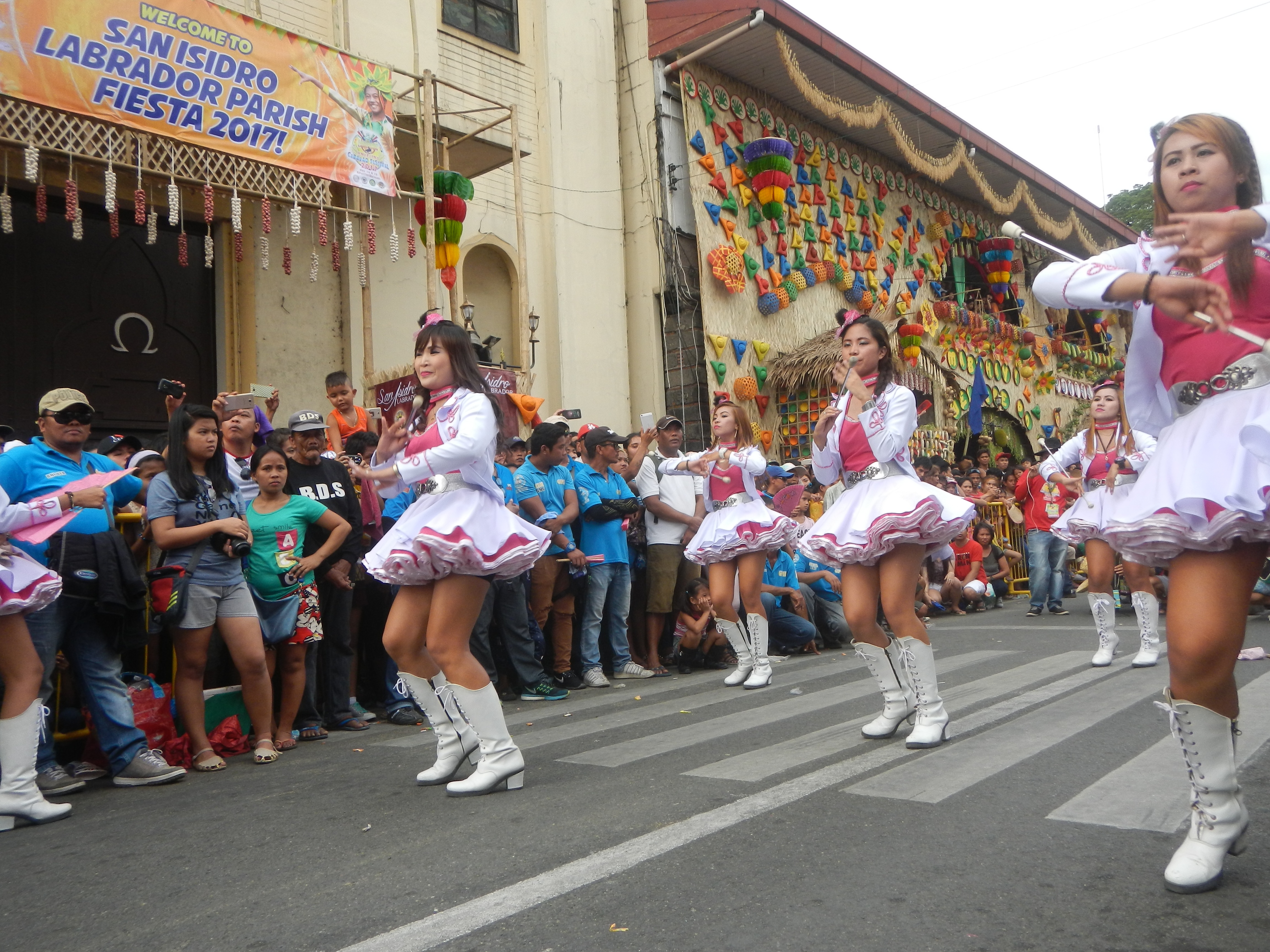 Eine Gruppe von Frauen in pinken und weißen Outfits tanzt auf einer Straße, umgeben von einer Menge Schaulustiger, mit Gebäuden, Bäumen und einem klaren blauen Himmel im Hintergrund.