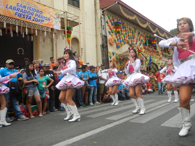 Eine Gruppe von Frauen in pinken und weißen Outfits tanzt auf einer Straße, umgeben von einer Menge Schaulustiger, mit Gebäuden, Bäumen und einem klaren blauen Himmel im Hintergrund.