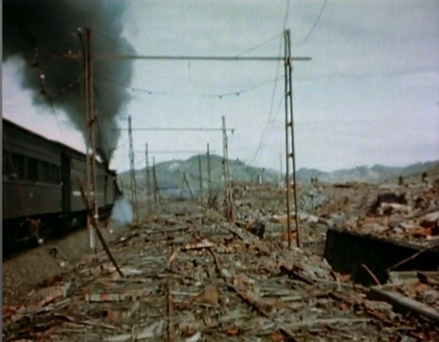 Ein Zug fährt durch eine verwüstete Landschaft mit Rauch, der aus ihm aufsteigt, umgeben von Strommasten und Drähten, mit Hügeln und einem klaren Himmel im Hintergrund und verstreuten Trümmern am Boden.