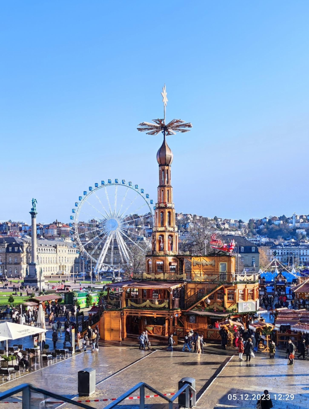 Ein belebter Stadtplatz mit einem zentralen Riesenrad, umgeben von Gebäuden, Bäumen und Menschen unter einem klaren blauen Himmel, mit Zelten und Absperrungen sichtbar.