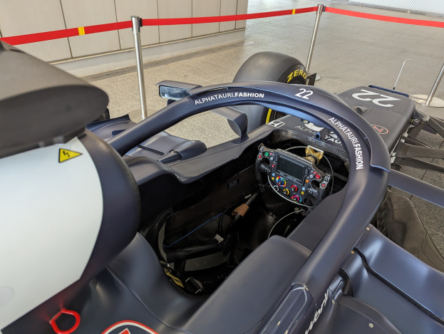 Cockpit eines Formel-E-Autos in einer Ausstellungshalle mit einem Lenkrad im Vordergrund, einer Wand im Hintergrund und dekorativen Bändern um die das Auto umgebenden Pfosten.