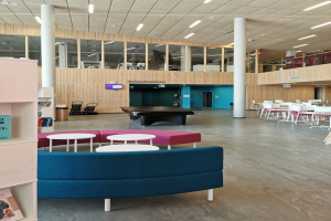 Ein geräumiger Raum mit zahlreichen Tischen, Stühlen, Sofas, Hockern, Bücherregalen, Säulen, Deckenleuchten und einem wandmontierten Fernseher, der an eine Schulbibliothek erinnert.