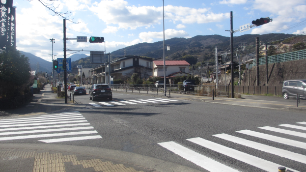Eine Stadtstraße mit Autos, einer Zebquerung, Strommasten, Verkehrsampeln, Gebäuden, Bäumen, Hügeln und einem bewölkten Himmel.