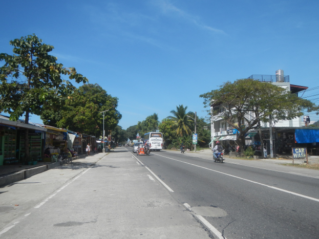 Eine belebte Stadtstraße mit Fahrzeugen, Fußgängern, Geschäftsfronten, Bäumen, Gebäuden, Strommasten und Überlandleitungen.