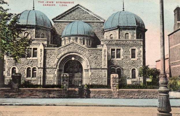 Alte Postkarte der jüdischen Synagoge in Cardiff, Ontario, Kanada, zeigend ein Gebäude mit Fenstern, Türen und Bögen, umgeben von Pflanzen, einem Zaun, Bäumen, einem Pfahl und einem bewölkten Himmel, mit Text oben.