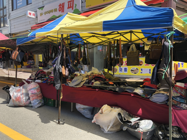 Ein Straßenmarkt mit Zelten, die verschiedene Gegenstände wie Taschen und Hüte anbieten, vor Gebäuden mit Fenstern und Texttafeln im Hintergrund.