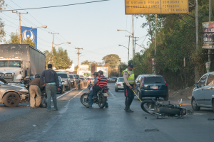 Eine Gruppe von Menschen um ein verunglücktes Motorrad auf der Straße herumstehend mit mehreren Fahrzeugen, darunter ein Lastwagen, und einem Hintergrund aus Bäumen, Pfosten, Lampen und Schildern unter dem Himmel.