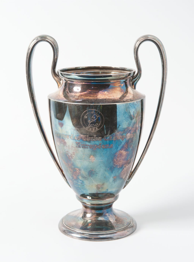 Der UEFA Champions League Pokal, ein stählerner Becher mit zwei Griffen und einem blauen und silbernen Farbschema, mit der Inschrift "UEFA Champions League" auf einem weißen Hintergrund.