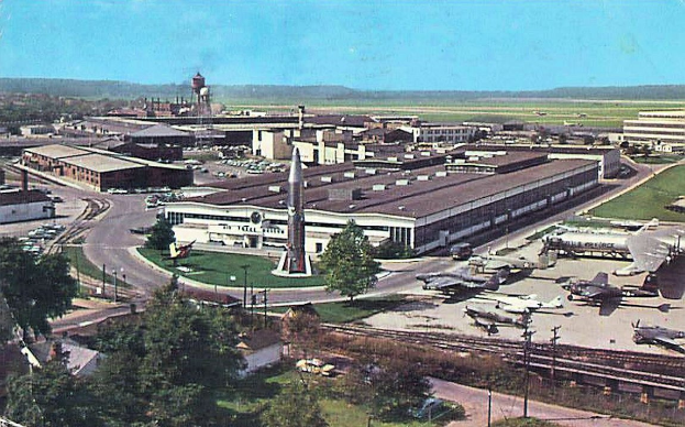 Alte Schwarz-Weiß-Flughafenszene mit geparkten Propellerflugzeugen, Terminalgebäuden, Bäumen, Straßen, Fahrzeugen, Strommasten, Gras, Bahnschienen, fernen Türmen, rollenden Hügeln und einem klaren Himmel, mit gedrucktem Text am unteren Rand.