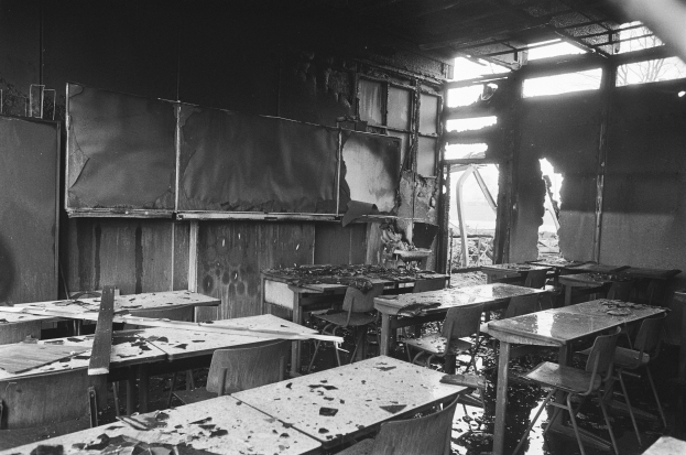 Schwarz-weißes Foto eines Klassenzimmers mit verbrannten Wänden, zerbrochenen Fenstern und leeren Tischen und Stühlen.