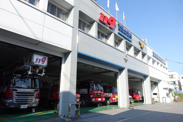 Feuerwache mit mehreren Feuerwehrautos vor der Tür, mit Fenstern, Säulen und Namensschildern, sowie Flaggen im Vordergrund, anderen Gebäuden im Hintergrund und einem klaren blauen Himmel.
