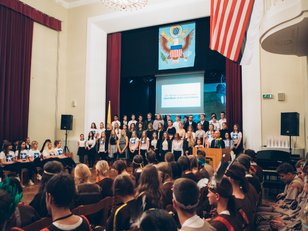 Eine Gruppe von Menschen steht vor einem sitzenden Publikum an einem Podium mit Mikrofon, flankiert von Fahnen und Lautsprechern, während eines 2. Kunst- und Kulturwochen-Events.