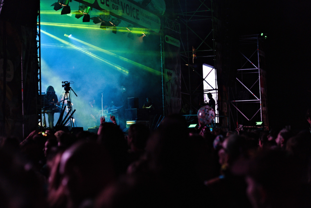 Eine Menge schaut einer Band zu, die auf einer hell erleuchteten Bühne mit Instrumenten, Mikrofonen und einem Banner spielt.