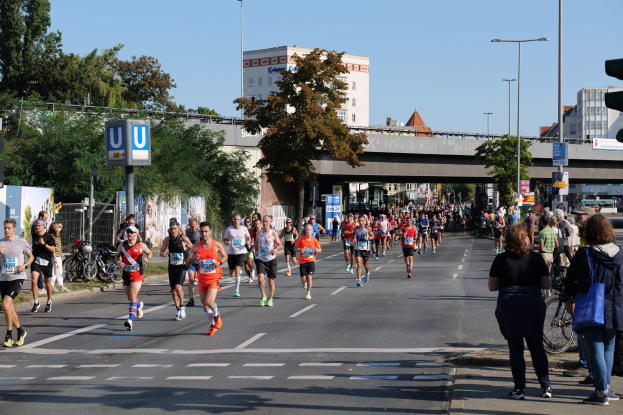 Eine Gruppe von Menschen, die bei einem Marathon auf einer von Bäumen gesäumten Straße mit Laternenpfählen, Schildern, Fahrrädern, einem Zaun, Gras, einer Brücke, Gebäuden und einem klaren blauen Himmel laufen.