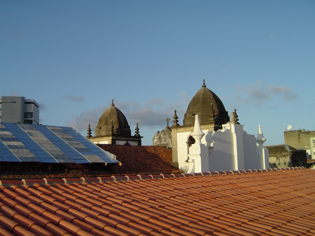 Stadtpanorama mit Gebäuden im Vordergrund, Solarpanels auf einem Dach und einem blauen Himmel im Hintergrund.
