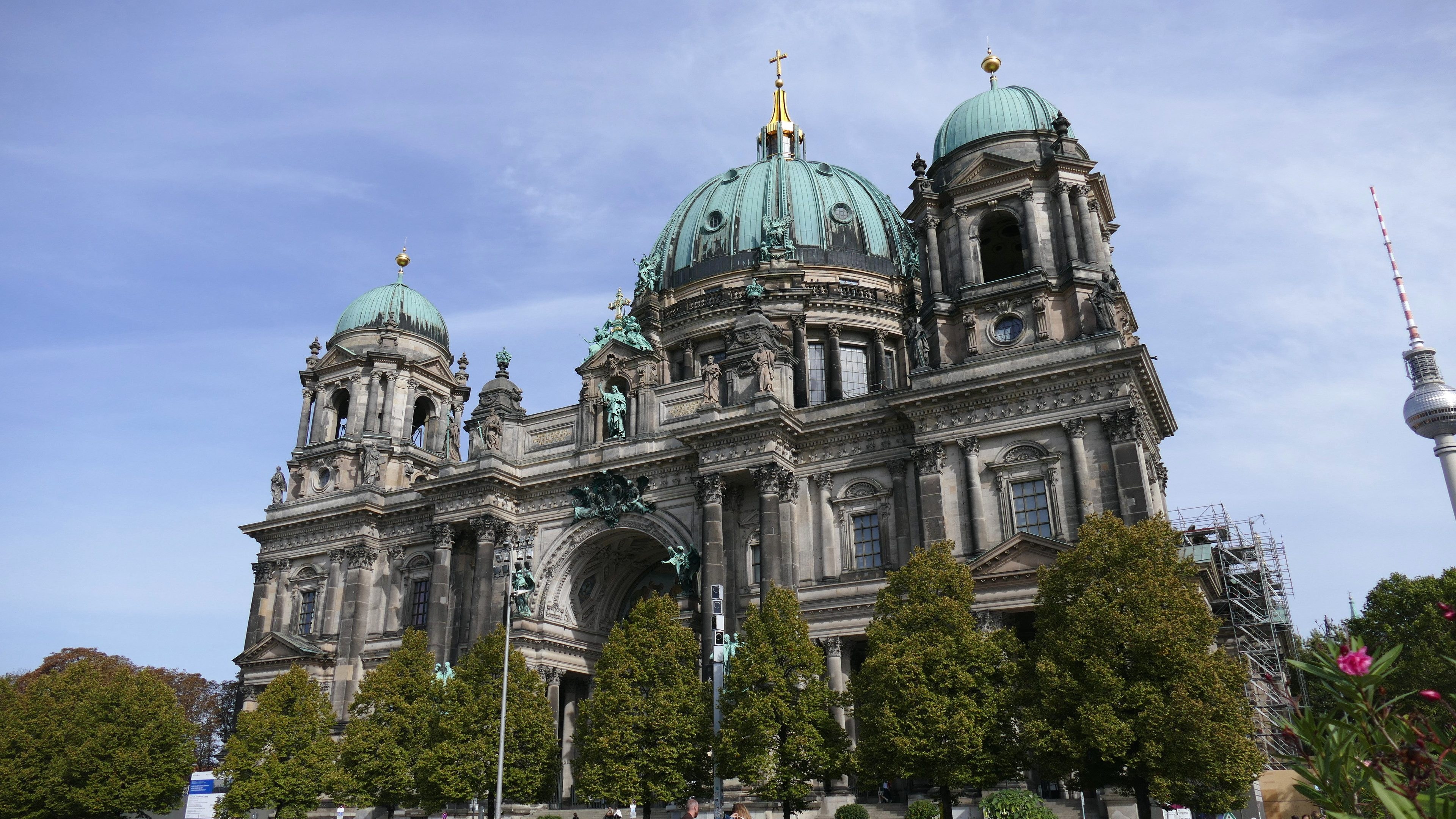 Der Berliner Dom, eine große Kathedrale mit Fenstern, Säulen, Bögen und Statuen, steht prominent in Berlin, Deutschland, mit Menschen in der Nähe und Bäumen, Blumenpflanzen und einem Turm im Vordergrund unter einem bewölkten Himmel.