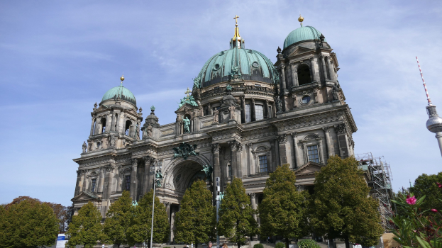 Der Berliner Dom, eine große Kathedrale mit Fenstern, Säulen, Bögen und Statuen, steht prominent in Berlin, Deutschland, mit Menschen in der Nähe und Bäumen, Blumenpflanzen und einem Turm im Vordergrund unter einem bewölkten Himmel.