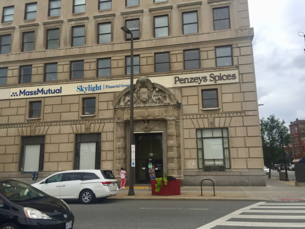 Großes Gebäude mit "Massachusetts Mutual Financial Group" Beschriftung, parkende Autos davor, Straßenlaternen, fahrende Fahrzeuge, Fußgänger auf dem Gehweg, Grünfläche und bewölkter Himmel.