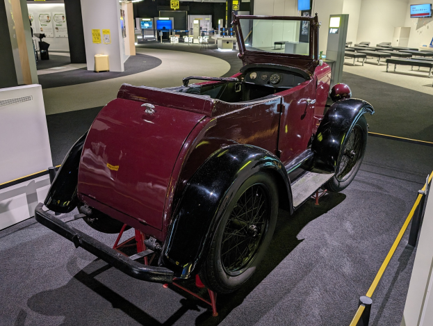 Ein rotes Oldtimer-Auto in einem Museum mit Informationsschildern, Barrieren und Beleuchtung ausgestellt.