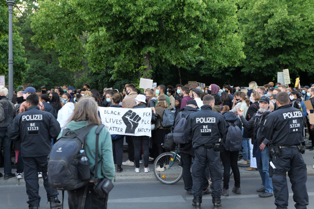 Eine große Gruppe von Menschen bei einer Black Lives Matter Demonstration in Berlin, einige halten Schilder und andere tragen Mützen und Taschen, mit einem Fahrrad im Vordergrund und Bäumen und einem Pfahl im Hintergrund.
