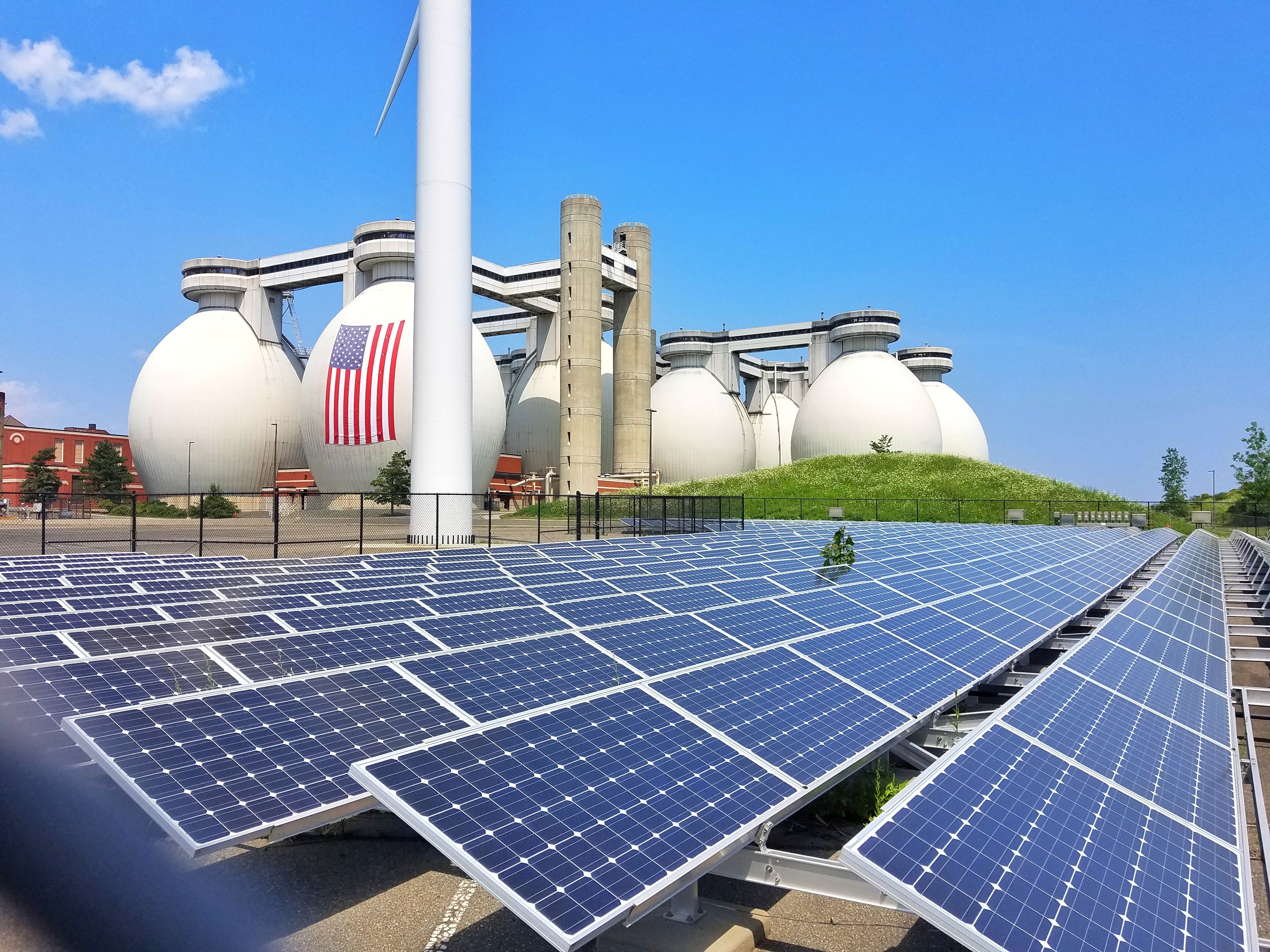 Solarpanels auf dem Dach eines Gebäudes mit einer Flagge, umgeben von einem Zaun und Grünflächen, Masten und anderen Strukturen, mit Gebäuden und einem bewölkten Himmel im Hintergrund.