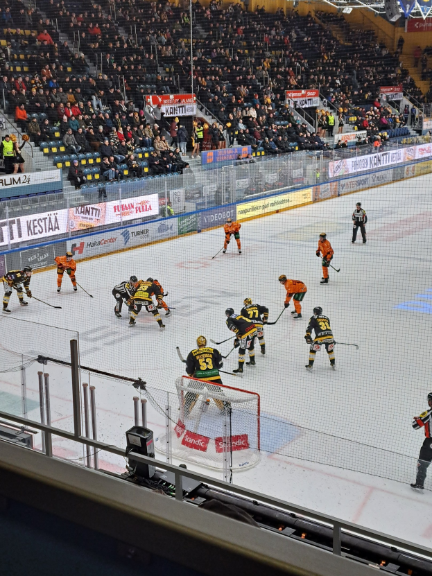 Eine Gruppe von Menschen, die Hockey auf einem Eisstadion spielt, mit einem Tor, einer Umzäunung, sitzenden Zuschauern, Bannern und Scheinwerfern.