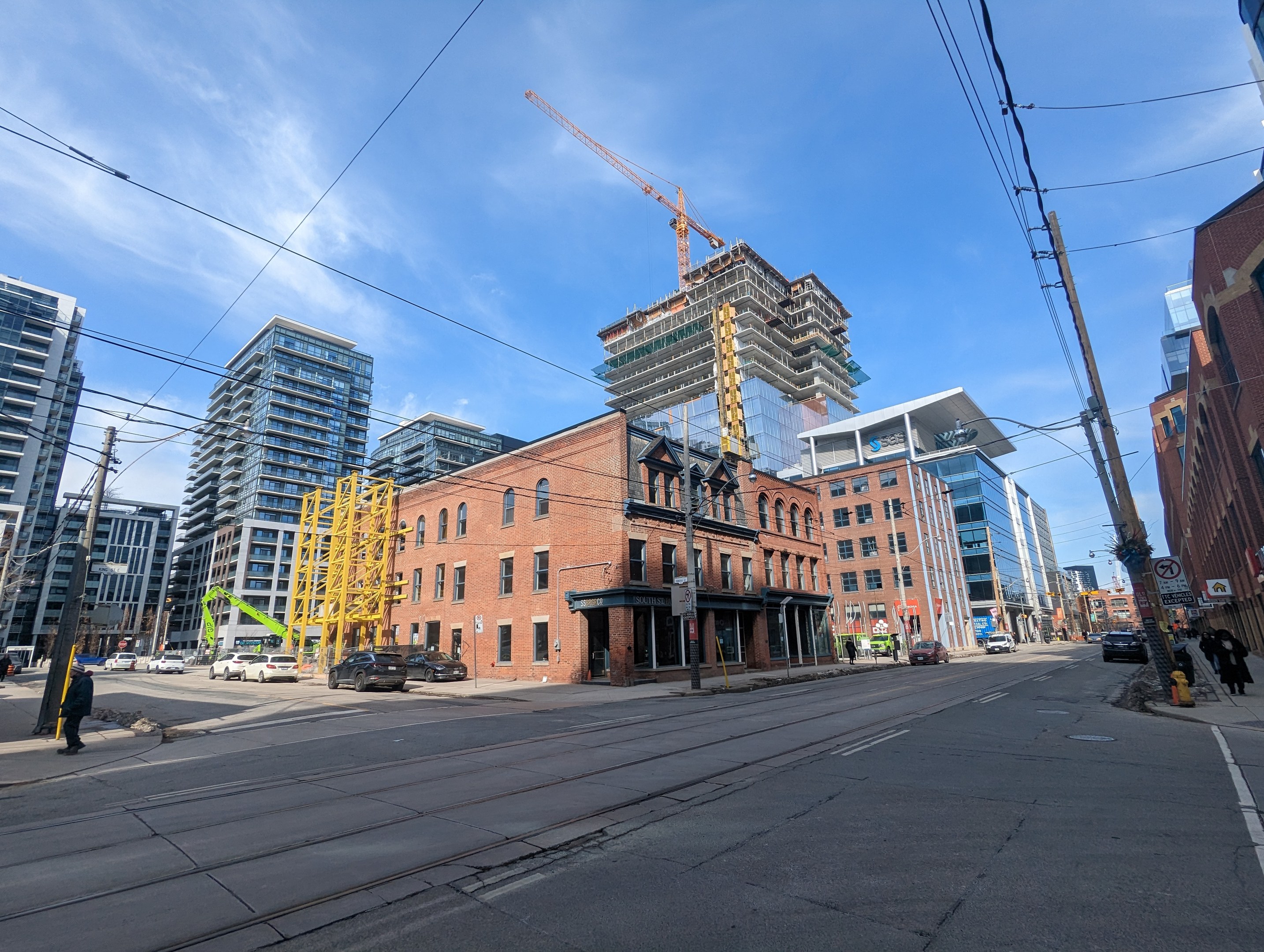 Eine belebte Stadtstraße in Toronto mit Fahrzeugen, Fußgängern, Strommasten, Schildern, Gebäuden und einer Baustelle im Hintergrund unter einem bewölkten Himmel.