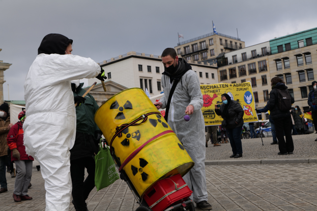 Eine Gruppe von Menschen in weißen Anzügen und Masken steht um einen gelben Behälter mit einem Wagen im Vordergrund, mit Schildern in der Hand, vor Gebäuden mit Fenstern und Fahnen auf Stangen unter einem bewölkten Himmel.