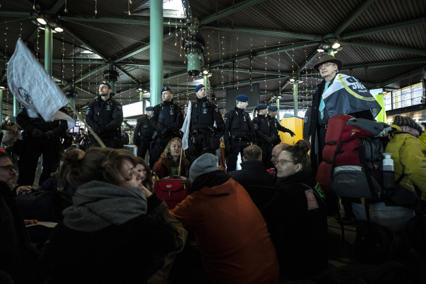 Eine Gruppe von Menschen vor einer protestierenden Menge an einem Bahnhof mit Säulen und Deckenbeleuchtung, einige tragen Mützen und tragen Taschen, andere halten Schilder und Transparente.