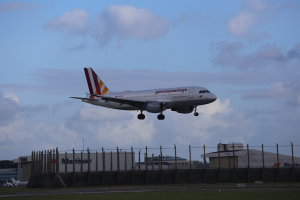 Germanwings Airbus A320-200 beim Start vom Frankfurt Airport, mit Gras darunter, einem Zaun im Vordergrund, Gebäuden und Bäumen im Hintergrund und einer bewölkten Himmel