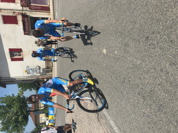 Gruppe von Radfahrern, die die Straße entlangfahren, an einem Gebäude mit Fenstern und Texttafeln vorbei, mit Bäumen und einem klaren blauen Himmel im Hintergrund und Zuschauern auf der Straße darunter.