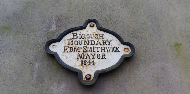 Metallplatte an der Seite eines Gebäudes, die die Borough-Grenze markiert, mit "Borough Boundary Edm Smithwick Mayor" in fetten weißen Buchstaben auf schwarzem Hintergrund.