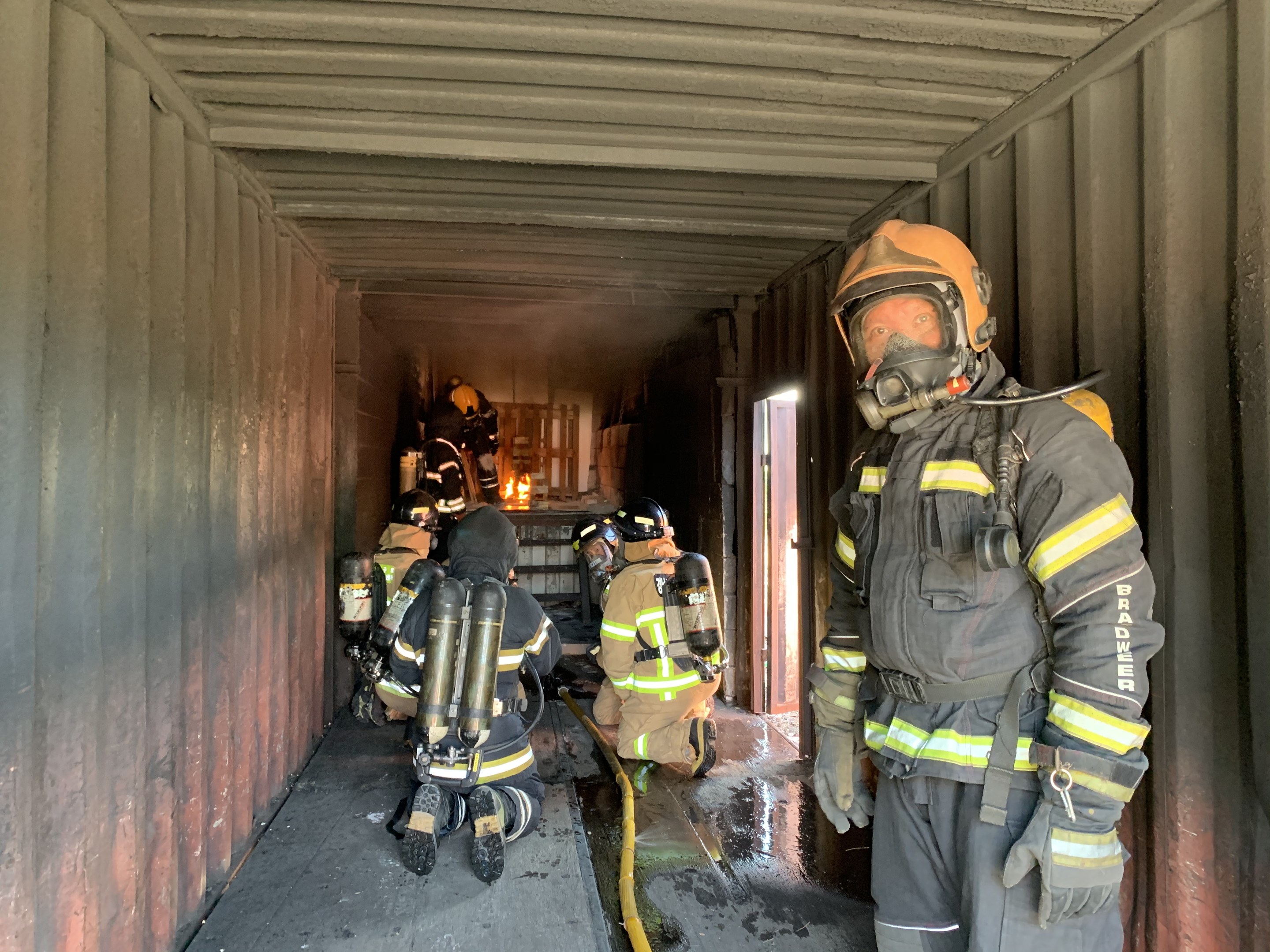 Feuerwehrleute in Schutzausrüstung stehen in einem Container mit einem Rohr auf dem Boden und einer Tür im Hintergrund.