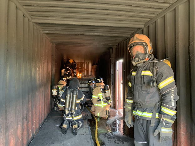 Feuerwehrleute in Schutzausrüstung stehen in einem Container mit einem Rohr auf dem Boden und einer Tür im Hintergrund.