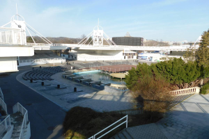Ein Blick auf das Olympische Stadion von einem hohen Punkt aus, mit verschiedenen Objekten und Grün im Vordergrund und einem bewölkten Himmel im Hintergrund.